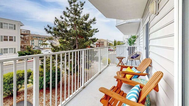 A balcony with wooden chairs and a small table, a white railing, and a view of neighboring houses and trees. - Newly Updated Condo Steps from the Beach w/ Balcony, Shared Rooftop Pool (Ocean City)