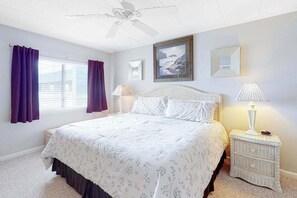 Une chambre à coucher comprenant un lit, une table de chevet, un ventilateur de plafond, une fenêtre avec des rideaux et un tableau encadré accroché au mur.