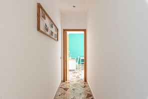 Um corredor estreito com chão estampado, uma porta de madeira que dá para um quarto com parede verde, e um quadro pendurado na parede.