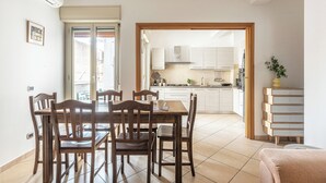 Un comedor con mesa y sillas de madera, una cocina al fondo y un gran ventanal corredizo que da al exterior.