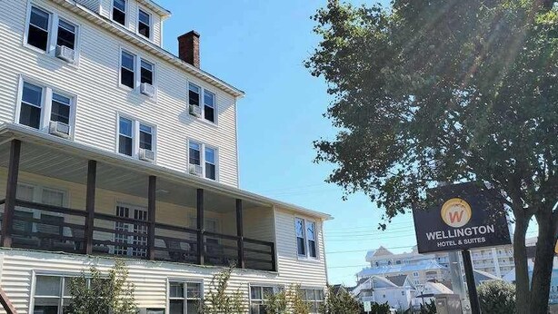 A two-story hotel with a balcony on the ground floor, a sign indicating it is Wellington Hotel & Suites, and a clear sky in the background. - Gorgeous & roomy condo with partial AC & full kitchen - steps from beach access (Ocean City)