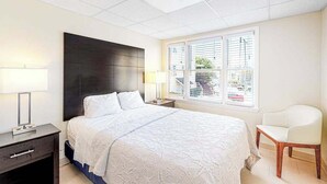 A bedroom with a bed, bedside lamps, a chair, a nightstand, and a window with blinds. - Gorgeous & roomy condo with partial AC & full kitchen - steps from beach access (Ocean City)