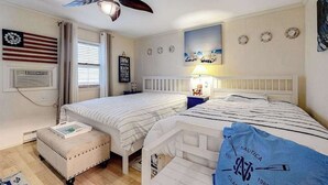 Een kamer met twee bedden, een plafondventilator, een vlag, een schilderij met een strandthema en een marine kussen.