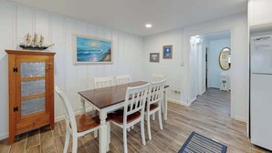 A dining area with a wooden table and chairs, a cabinet with glass doors, a painting of a seascape, and a hallway leading to another room. - Newly remodeled, ocean block condo with balcony, central AC & W/D (Ocean City)