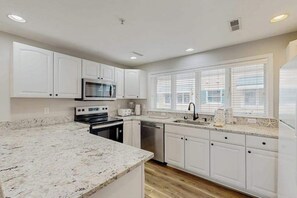 A modern kitchen with white cabinets, granite countertops, a microwave, stove, dishwasher, and a window with blinds. - Newly Renovated, 3-Story Townhome with Balconies, WiFi, AC & Washer/Dryer (Ocean City)