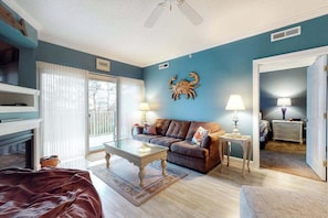 A living room with a sofa, coffee table, and lamps. There is a fireplace, a door leading to another room, and a ceiling fan. - Summer House 102 (Ocean City)