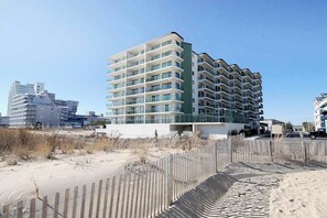 A multi-story hotel with balconies, located near a sandy beach with a wooden fence. - Side Ocean View Condo near the Boardwalk with Balcony, AC, WiFi & Shared Pool (Ocean City)