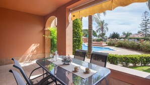 A glass table with chairs, a small ship model, and two cups on a patio with a view of a swimming pool and greenery.