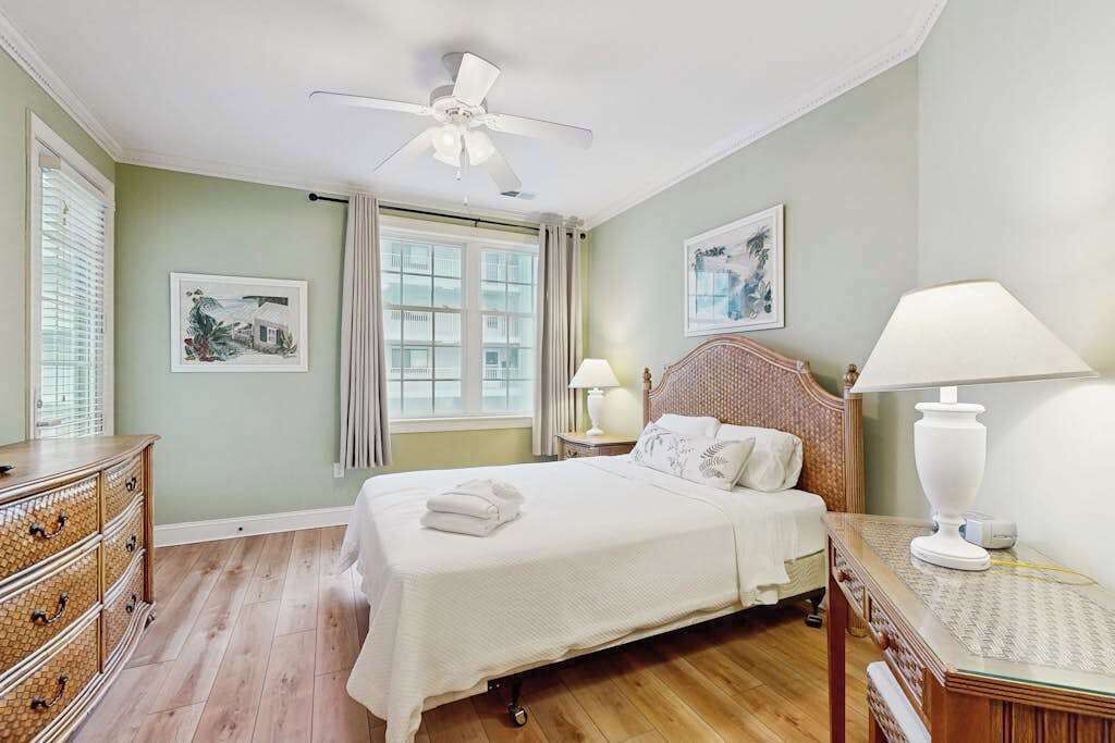 A hotel room with a bed, a ceiling fan, a nightstand with a lamp, a dresser, and two framed pictures on the wall.
