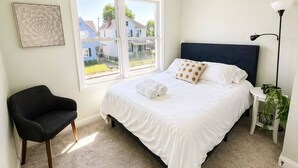 A bedroom with a bed, a chair, a nightstand, a lamp, and a window with a view of houses. - Sleeps 10 •4BR Home •Perfect for Groups & Families (Columbus)