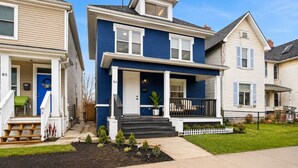 A two-story residential building with a front porch, a small garden, and a white picket fence. - Sleeps 10 •4BR Home •Perfect for Groups & Families (Columbus)