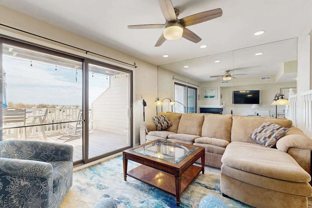 A spacious living room with a large glass coffee table, a beige sofa, a patterned armchair, and a ceiling fan.