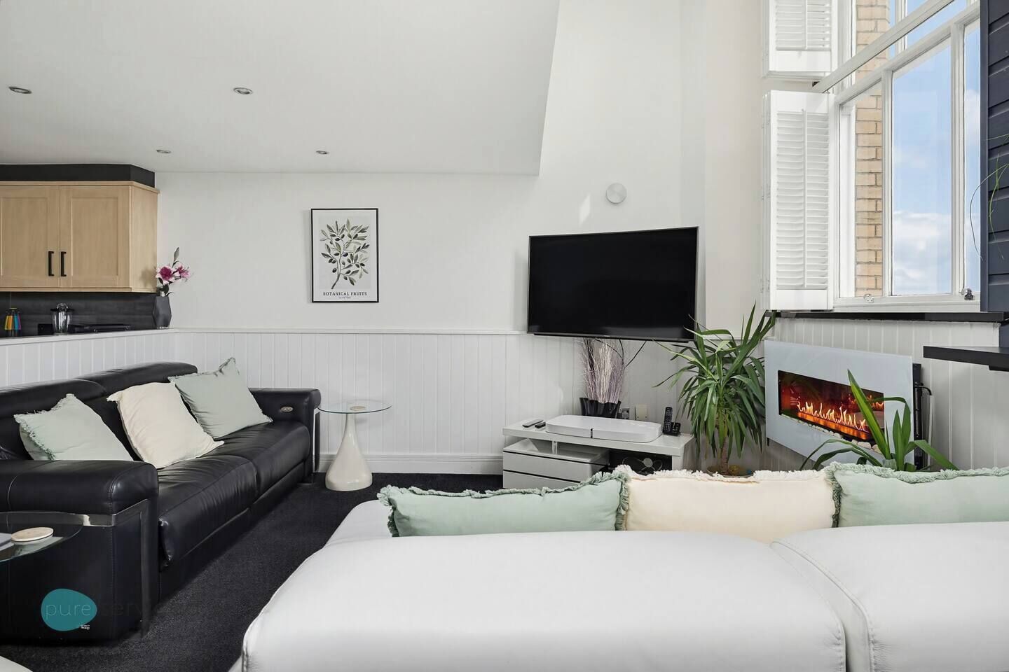 A modern living room with a black leather sofa, a white armchair, a glass coffee table, a flat-screen TV, a fireplace, and a kitchen area with wooden cabinets.