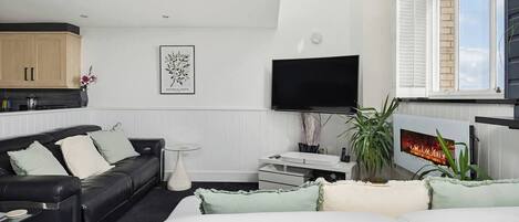 A modern living room with a black leather sofa, a white armchair, a glass coffee table, a flat-screen TV, a fireplace, and a kitchen area with wooden cabinets.
