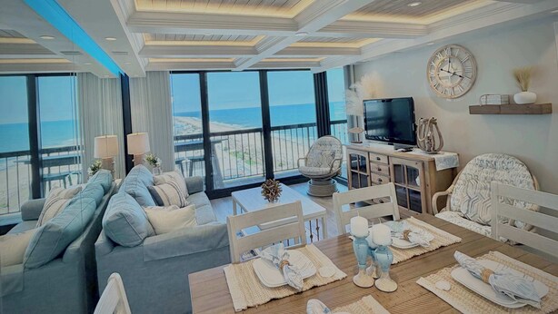 Ein modernes Wohnzimmer mit großem Fenster mit Blick auf den Strand, ein Essbereich mit Holztisch für vier Personen und ein Fernseher auf einem Holzschrank.