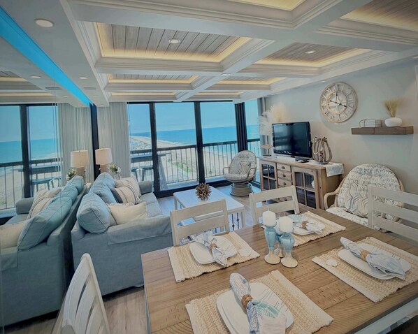 Ein modernes Wohnzimmer mit großem Fenster mit Blick auf den Strand, ein Essbereich mit Holztisch für vier Personen und ein Fernseher auf einem Holzschrank.