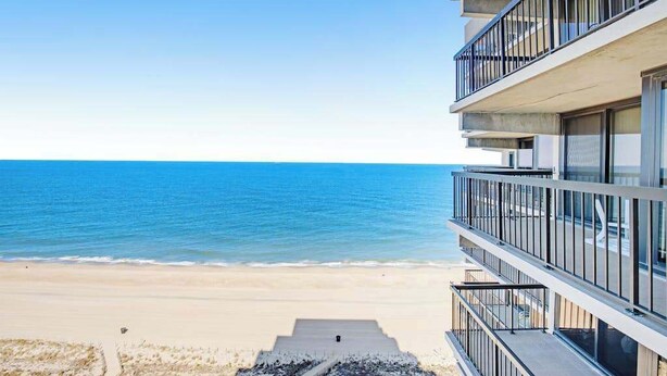 Un hôtel situé en bord de mer, offrant une vue sur l’océan, doté de balcons et d’un chemin en béton menant à la plage.