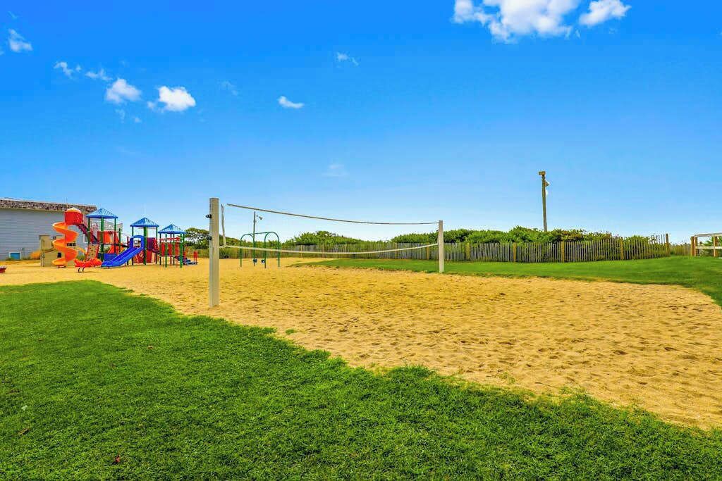 Un terrain de volleyball de sable avec un filet, entouré de pelouse verdoyante et un espace de jeux avec des toboggans.