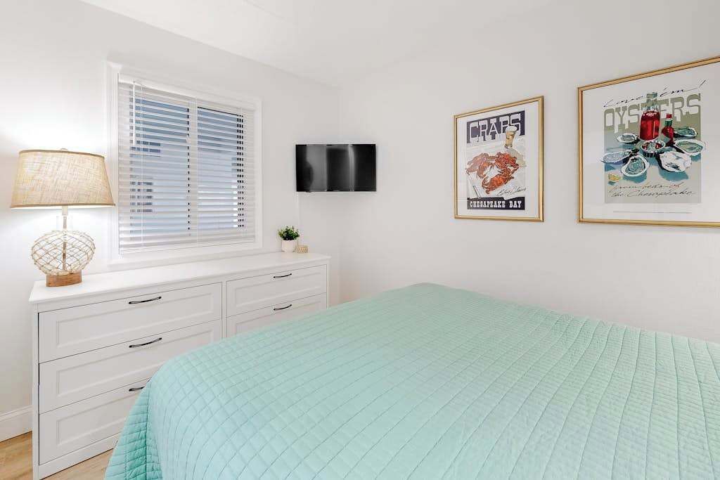A bedroom with a bed, a TV, a window with blinds, a lamp, and two framed pictures on the wall.