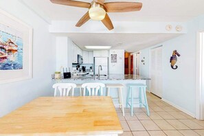 A dining area with a wooden table and chairs, a kitchen in the background, and a ceiling fan. - Stunning beach condo with balcony, views, pools, gym, game room & sauna (Ocean City)