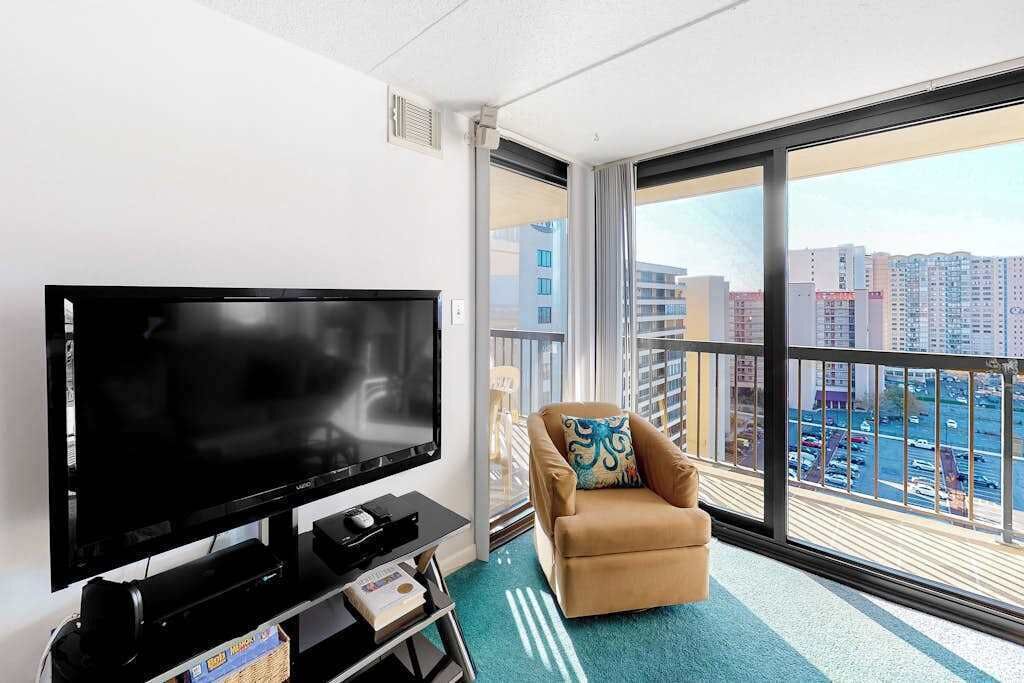 A modern hotel room with a large flat-screen TV, a comfortable armchair, and a balcony with a city view.