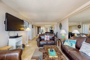 A living room with a flat-screen TV, leather sofas, a coffee table with a book, and a dining area in the background. - Modern beach condo with balcony, pools, gym, game room & sauna (Ocean City)