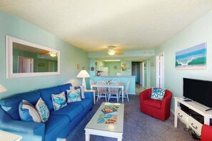 A living room with a blue sofa, a red armchair, a coffee table, and a TV stand. - Coastal condo with pools, game room, gym, sauna & central AC (Ocean City)