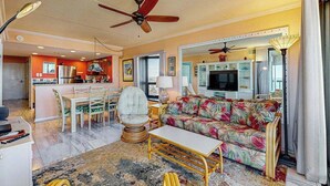 A living room with a sofa, a dining table, chairs, a ceiling fan, and a kitchen in the background. - Incredible beach condo with balcony, pools, game room, gym & sauna (Ocean City)