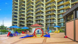 A multi-story residential building with balconies, a children's play area with slides and a ring, and outdoor seating with umbrellas. - Sunny beach condo with balcony, game room, pools, sauna & gym (Ocean City)