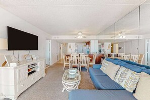 A modern living room with a sofa, a TV, a dining area with chairs, and a kitchen in the background. - Ocean-view condo on the beach with balcony, pools, saunas, tennis & gym (Ocean City)
