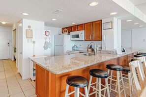 A kitchen with wooden cabinets, a granite countertop, a microwave, and a refrigerator. There are four bar stools at the counter. - Ocean-view condo on the beach with balcony, pools, saunas, tennis & gym (Ocean City)
