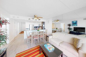 A modern living room with a sofa, a coffee table, a TV, and a dining area with chairs and a table. - Ocean-view condo with balcony, washer/dryer, pools, saunas, gym & tennis (Ocean City)