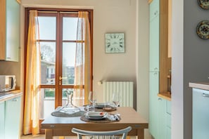 Een eethoek met een tafel gedekt voor twee personen, een raam met gordijnen, een wandklok en keukenkastjes.