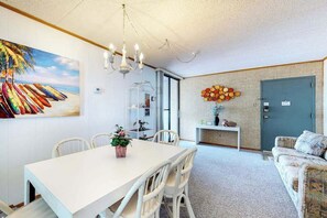 A dining area with a white table and chairs, a chandelier, a painting of surfboards, and a door with a keycard lock. - Oceanfront condo with two balconies, bay & ocean views, AC, WiFi, shared pool (Ocean City)