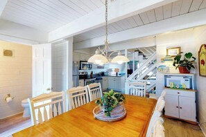A dining area with a wooden table, white chairs, a hanging light fixture, and a kitchen in the background. - Oceanfront Townhouse w/Private Balcony, Great Views, Washer/Dryer, Central AC (Ocean City)
