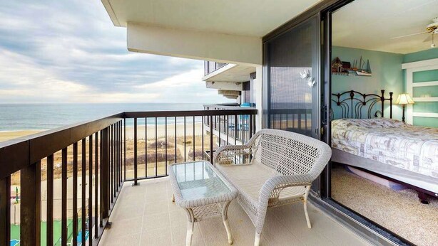 En balkon med et glastabel og en fletstol, en seng med et mønstret sengetæppe, og udsigt til stranden.