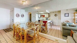 Un coin repas avec une table en verre et des chaises en osier, une cuisine à l’arrière-plan et un salon avec un canapé.
