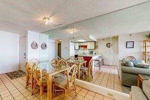 A dining area with a glass table and wicker chairs, a kitchen in the background, and a living area with a sofa. - Oceanfront Condo w/ Balcony, Ocean View, Central AC, W/D & Beach Access (Ocean City)