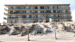 Un hôtel à plusieurs étages doté de balcons, situé sur une plage de sable fin, avec des dunes en premier plan.