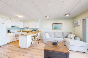A modern kitchen with white cabinets, a dining area with a wooden table, and a living room with a sofa and a painting on the wall. - Sandpiper Dunes 808 (Ocean City)