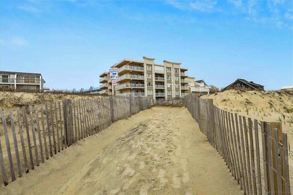 A beachfront hotel with a sandy walkway leading to the entrance, a wooden fence, and a clear blue sky.