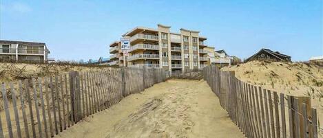 A beachfront hotel with a sandy walkway leading to the entrance, a wooden fence, and a clear blue sky.