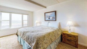 A bedroom with a bed, two bedside lamps, a nightstand, a framed picture on the wall, and a large window with a view.