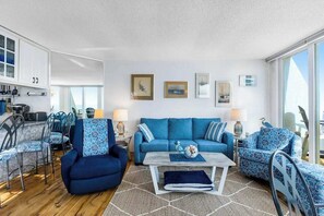 Ein modernes Wohnzimmer mit einem blauen Sofa, passenden Sesseln und einem Couchtisch. Es gibt eine Essecke mit Stühlen und einem Tisch sowie eine Küchenzeile mit Schränken und einem Herd.