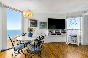 Ein Zimmer mit Esstisch, Stühlen, einem Fernseher und einem großen Fenster mit Meerblick.