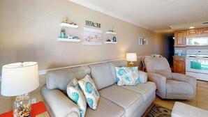 A living room with a beige sofa, a chair, a lamp, and wall decorations including a 'BEACH' sign. - Polynesian 309 (Ocean City)