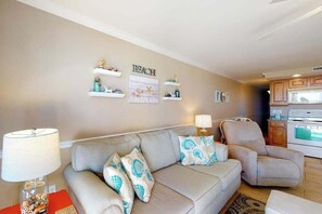 Una sala de estar con un sofá beige, una silla, una lámpara y adornos en la pared, incluyendo un cartel que dice 'BEACH'.