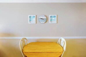 Un comedor con una mesa de madera y sillas blancas, una obra de arte enmarcada en la pared y un reloj.