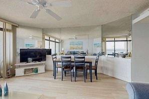 A modern living room with a TV, dining table, chairs, and a ceiling fan. - Bright, oceanfront condo with balcony, AC, laundry & outdoor pool (Ocean City)