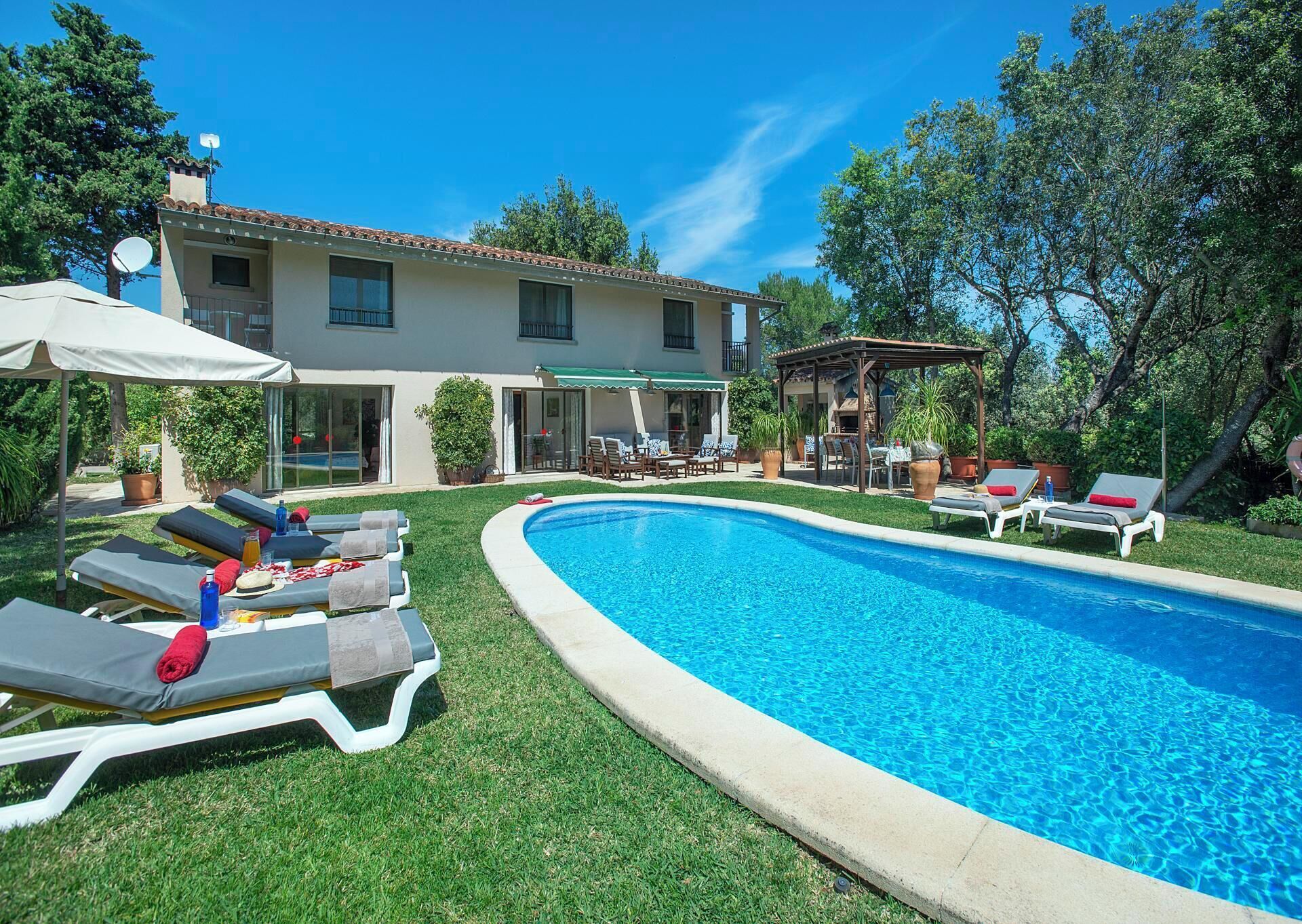 A swimming pool with lounge chairs and a pergola, surrounded by a well-maintained lawn and a house with a tiled roof.